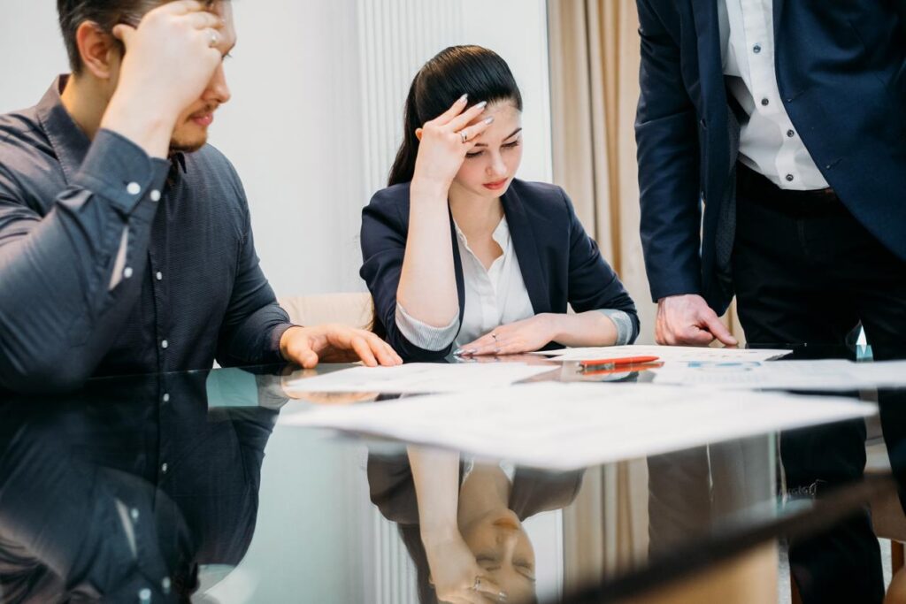 Man and woman sit at a table full of documents as they lean their heads on their hands in frustration